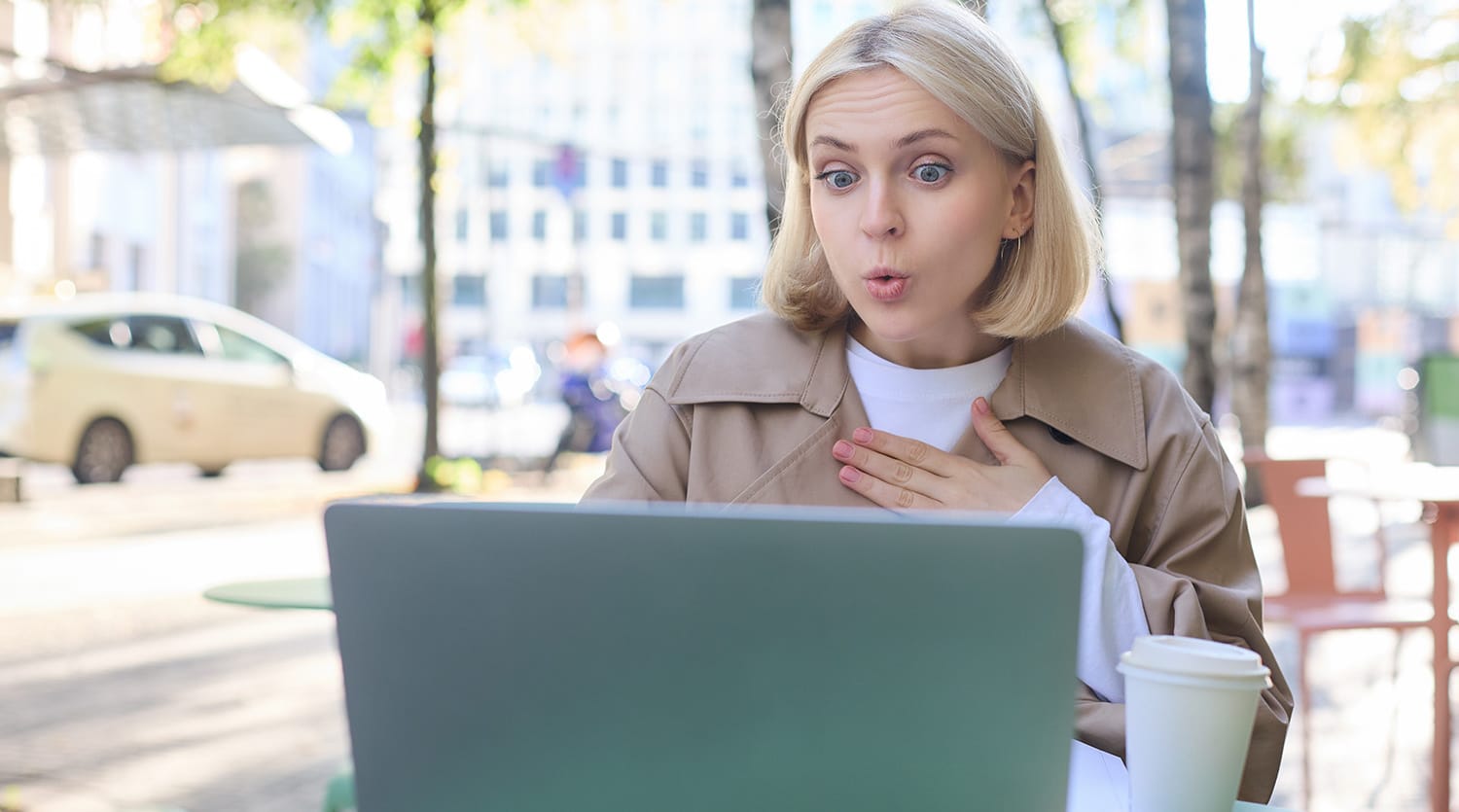 Close up portrait of young blond woman, sitting outdoors with laptop, drinking coffee in cafe, looking amazed and surprised at device screen, hears awesome news