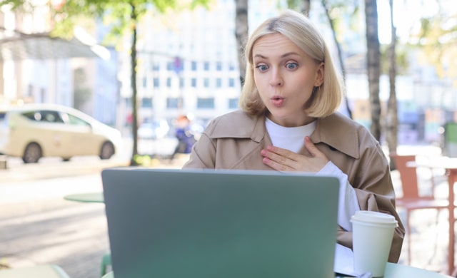 Close up portrait of young blond woman, sitting outdoors with laptop, drinking coffee in cafe, looking amazed and surprised at device screen, hears awesome news