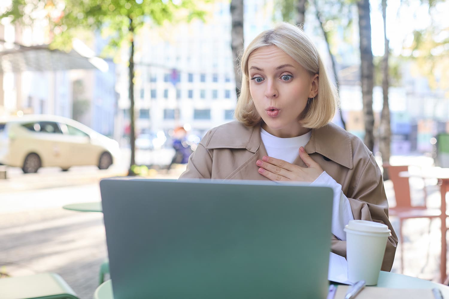 Close up portrait of young blond woman, sitting outdoors with laptop, drinking coffee in cafe, looking amazed and surprised at device screen, hears awesome news